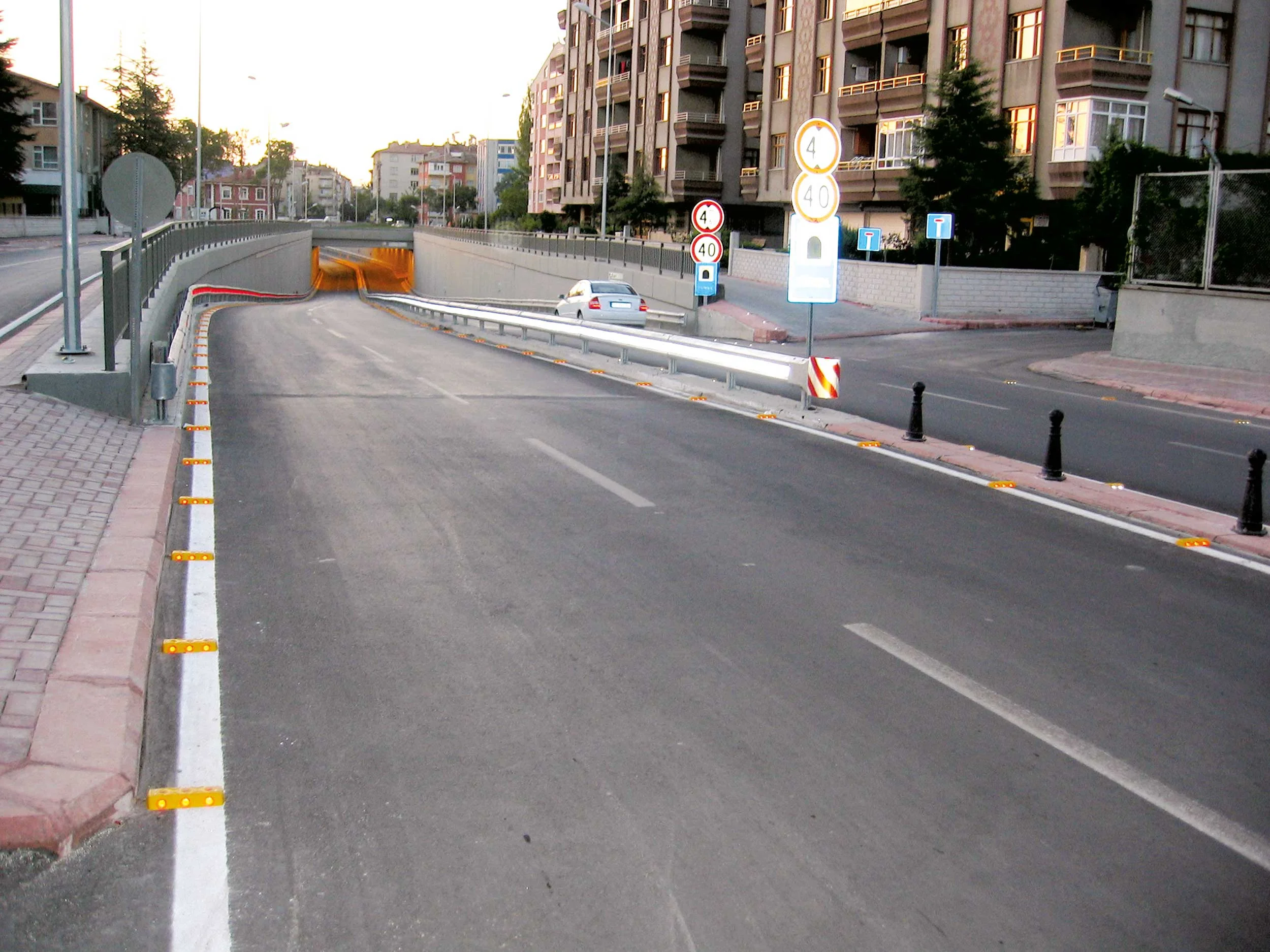 Straße mit Unterführung und weißen Leitpfosten mit orangenen Reflektoren entlang der Fahrbahn, Wohngebäude im Hintergrund