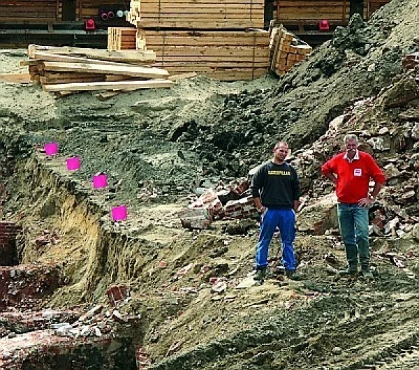 Zwei Männer stehen auf einer Baustelle mit Erdaushub, im Hintergrund Holzpaletten und Baumaterial