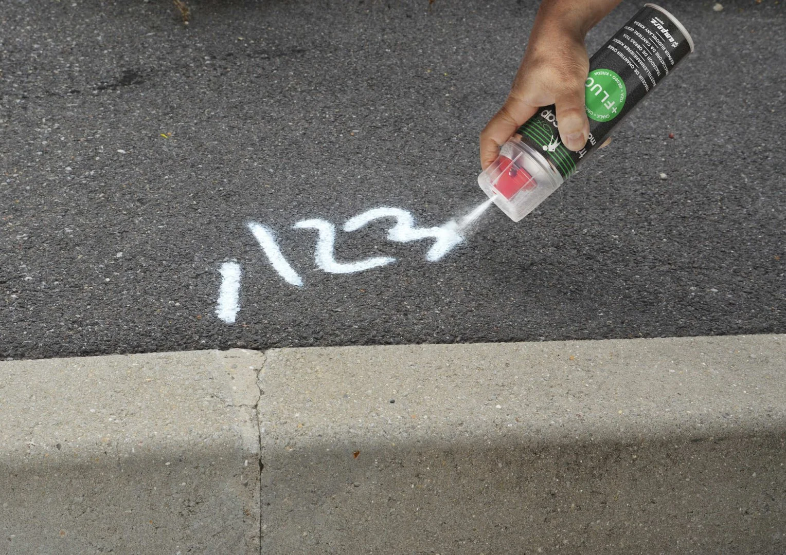 Hand hält Sprühflasche mit grünem Label und sprüht weiße Markierung in Form von Zahlen auf dunklen Asphalt neben Bordstein